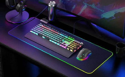 RGB backlit wireless gaming mouse with ergonomic honeycomb design on a large RGB mouse pad next to a mechanical keyboard in a gaming setup
