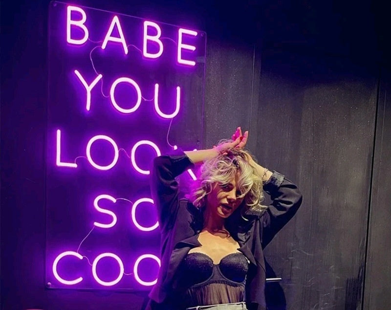A stylish woman poses in front of a neon LED light sign that says 'Babe You Look So Cool' in vibrant purple.